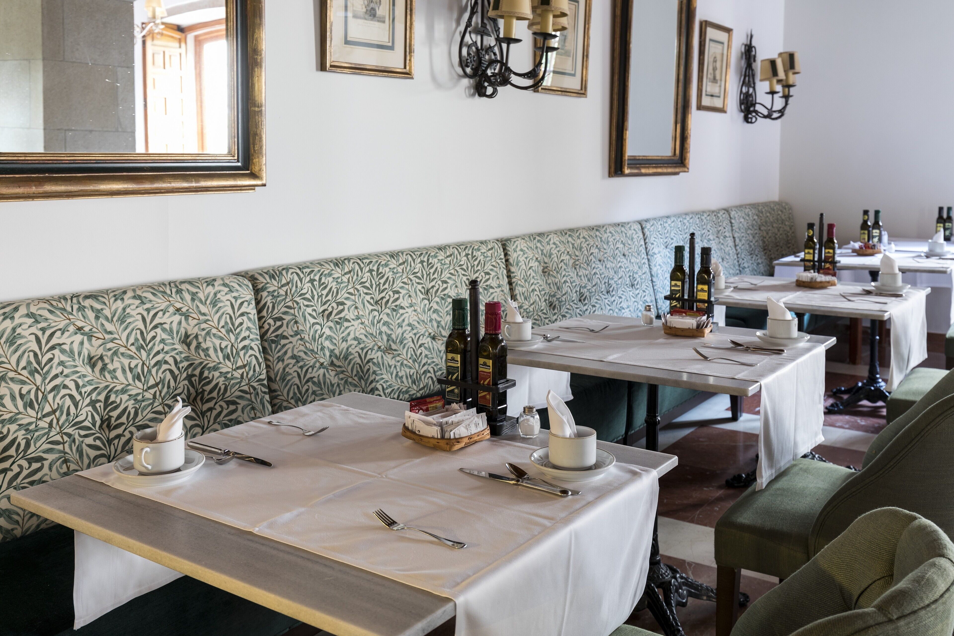 Un restaurante con una amplia zona de asientos tipo banco, mesas con manteles blancos y botellas de vino en cada una.