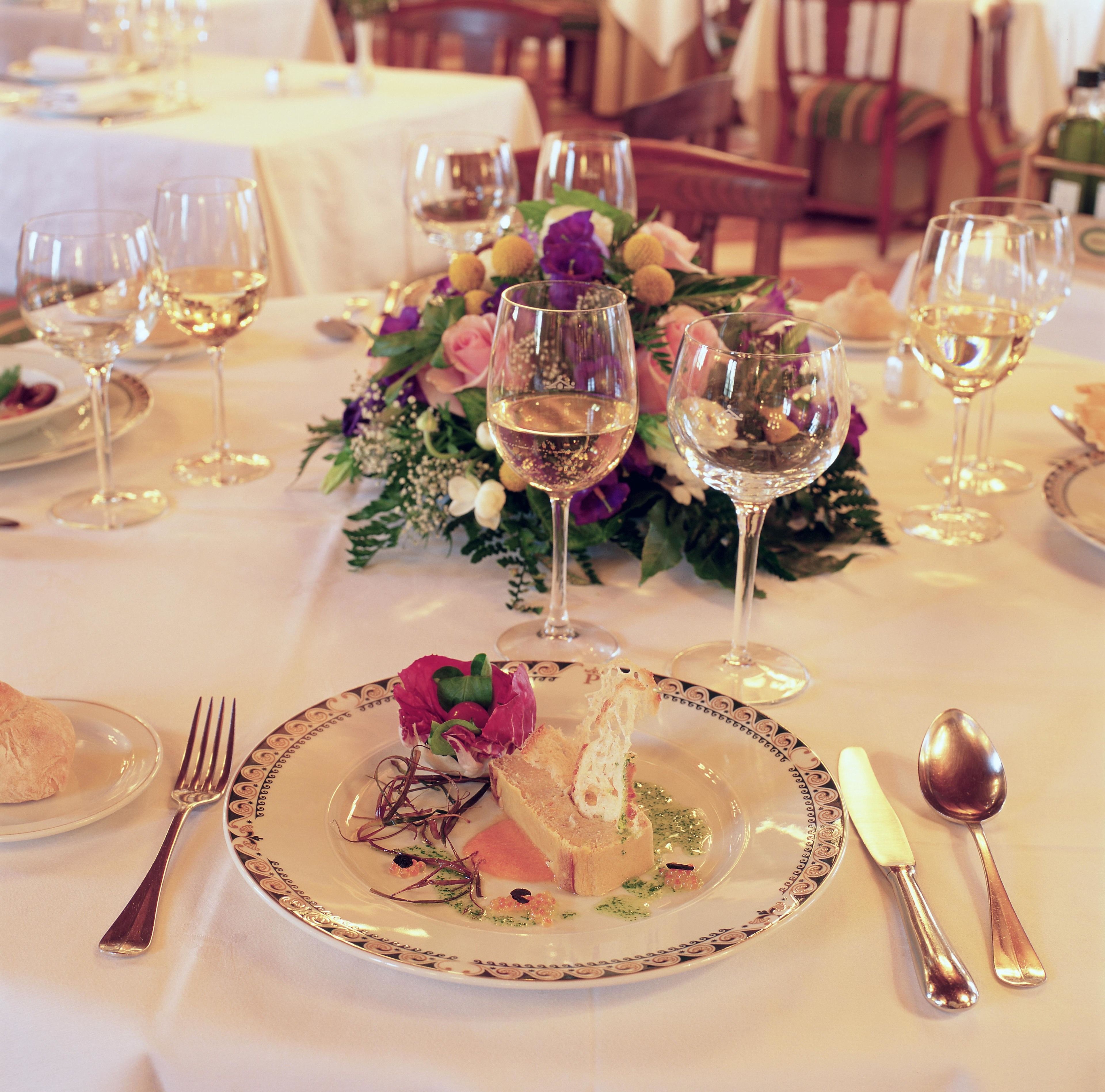 Mesa formal dispuesta con mantel blanco, copas de vino, platos con comida y cubiertos.