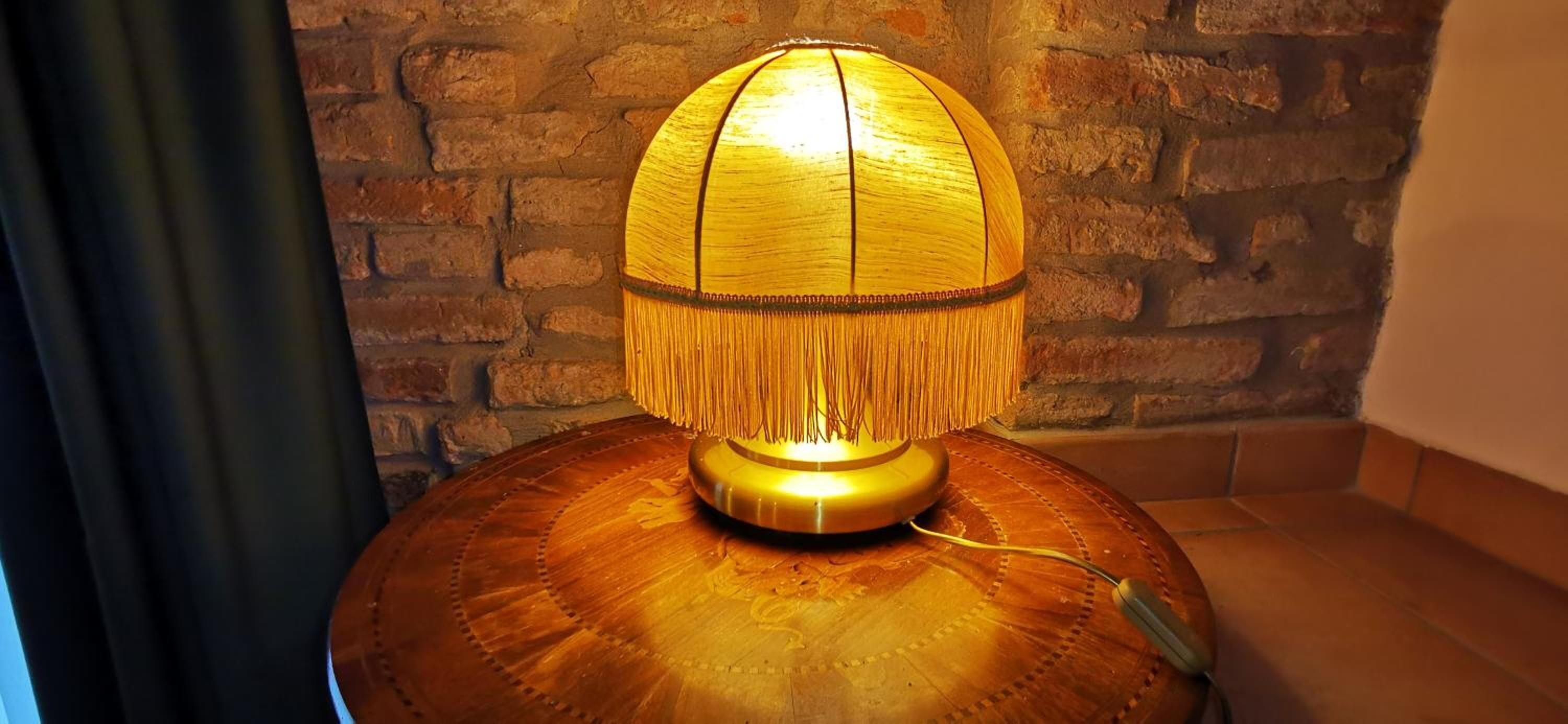 Eine Lampe mit Holzfuß und goldener Schirm mit Quasten auf einem runden Tisch vor einer Ziegelwand.