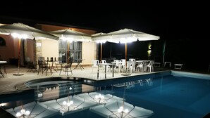 Una piscina con sillas de descanso y sombrillas, rodeada de mesas y sillas, que indica una zona de piscina.