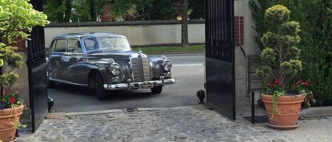 Ein klassisches Auto vor einem verschlossenen Eingangstor mit Kopfsteinpflaster und Kübelpflanzen.