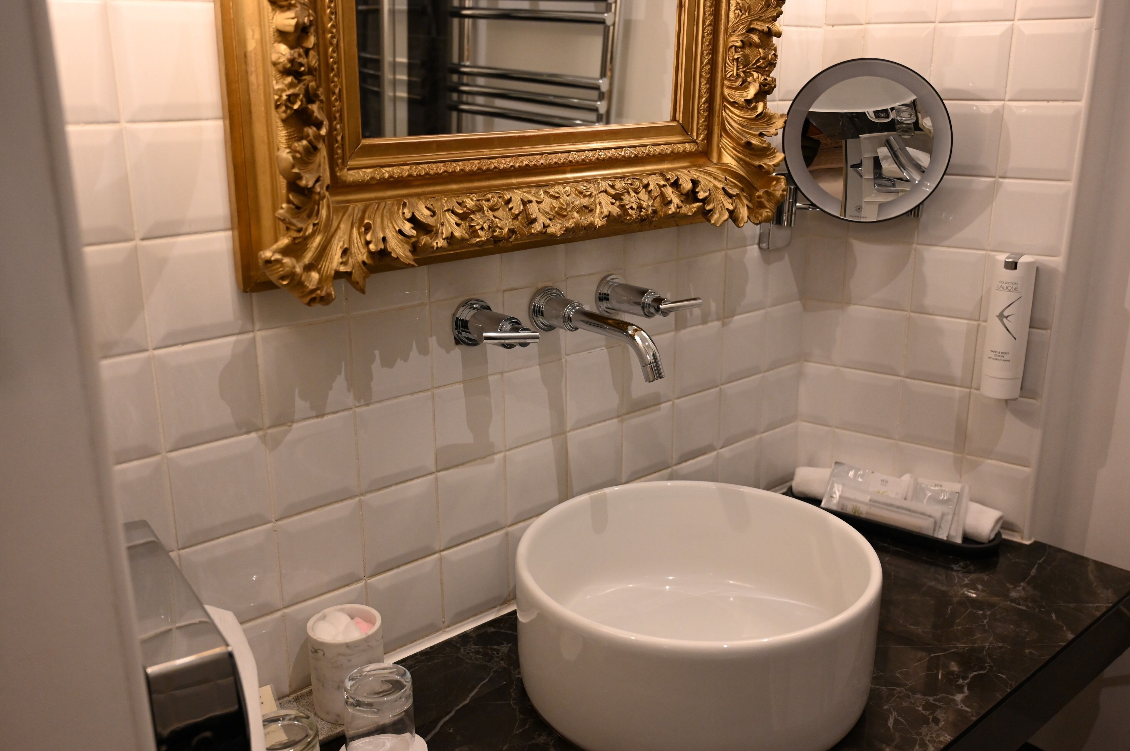 Une salle de bain avec un lavabo blanc, deux robinets, un miroir à cadre doré, un miroir rond et un distributeur de serviettes.