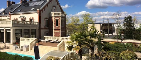 A large, historic building with a prominent tower, a swimming pool, and a well-maintained garden with various plants and a pergola.