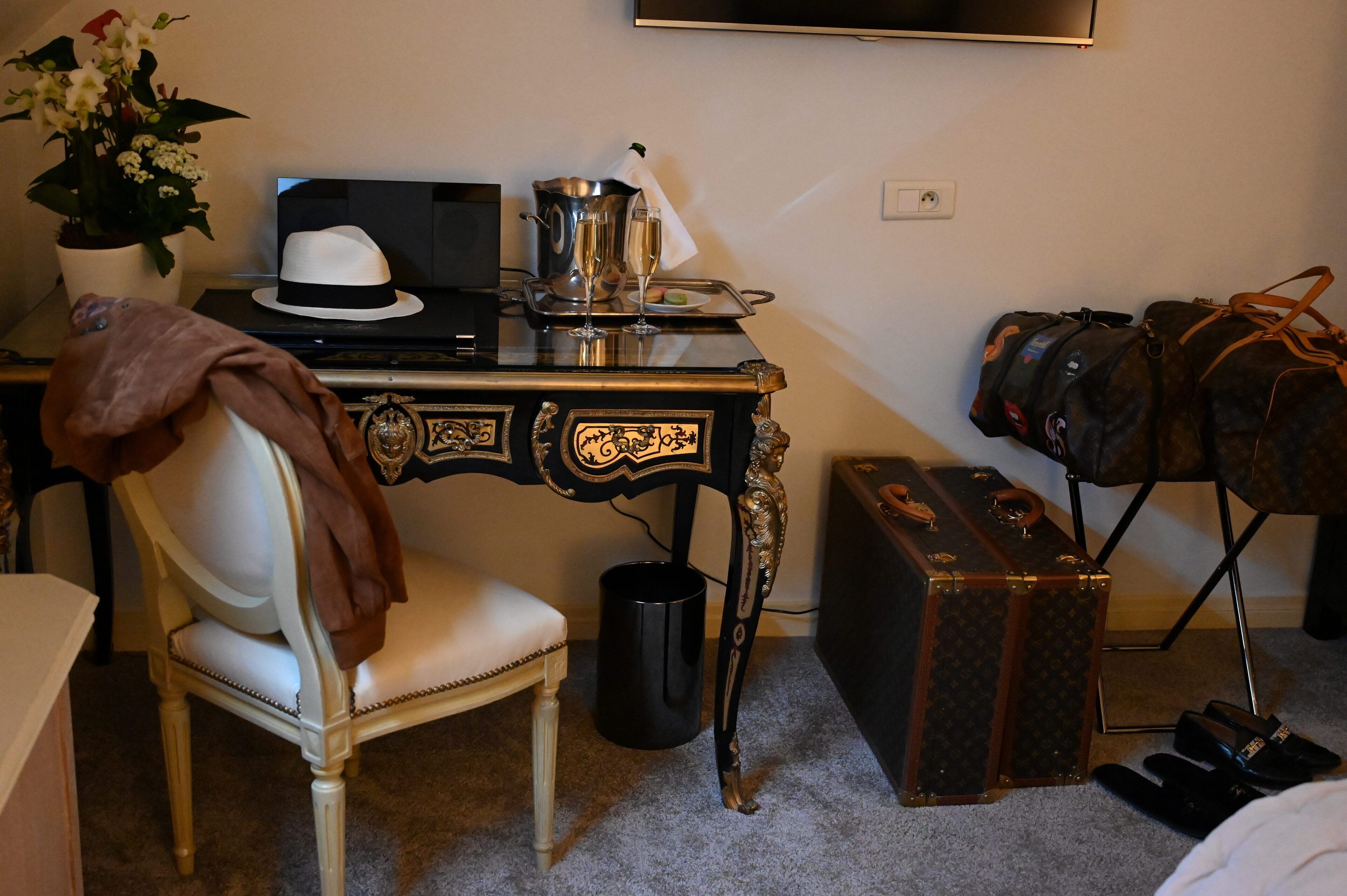 A hotel room with a vintage desk, a chair, a potted plant, a hat, a tray with glasses, a suitcase, and a duffel bag.