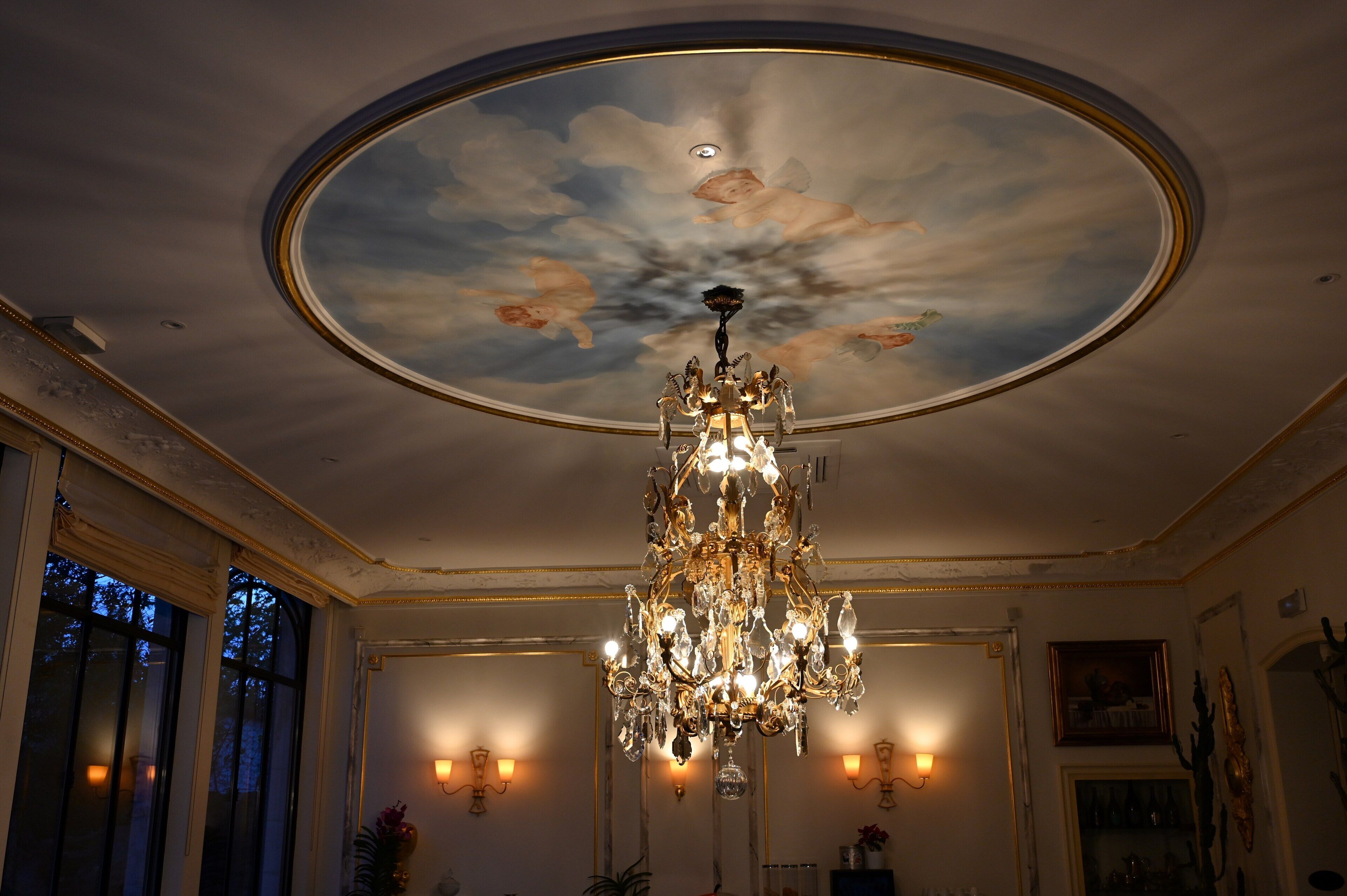 A grand room with a large chandelier, a ceiling mural, and wall sconces.