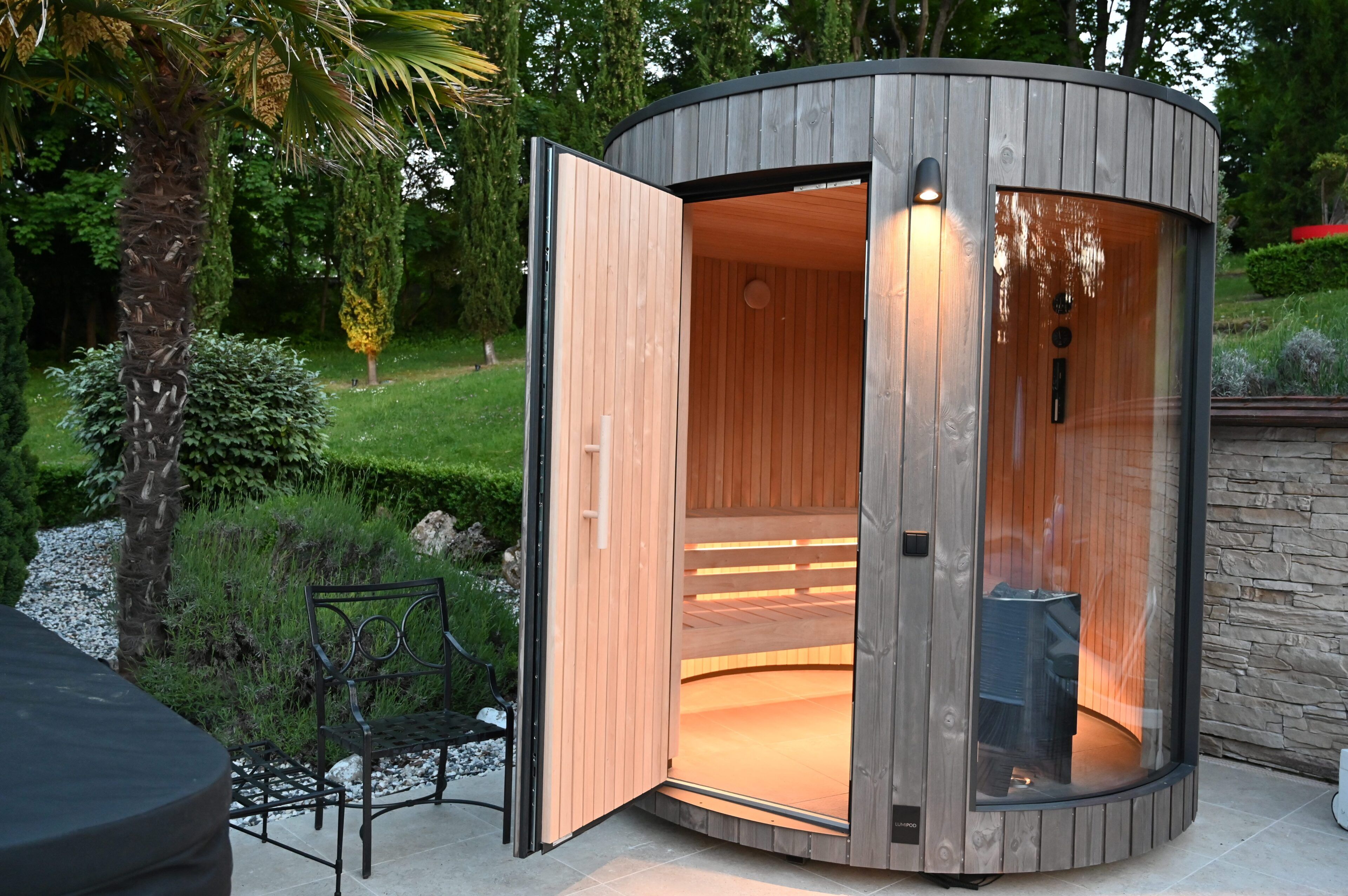 A circular wooden structure with an open door, a bench outside, and a stone wall in the background.