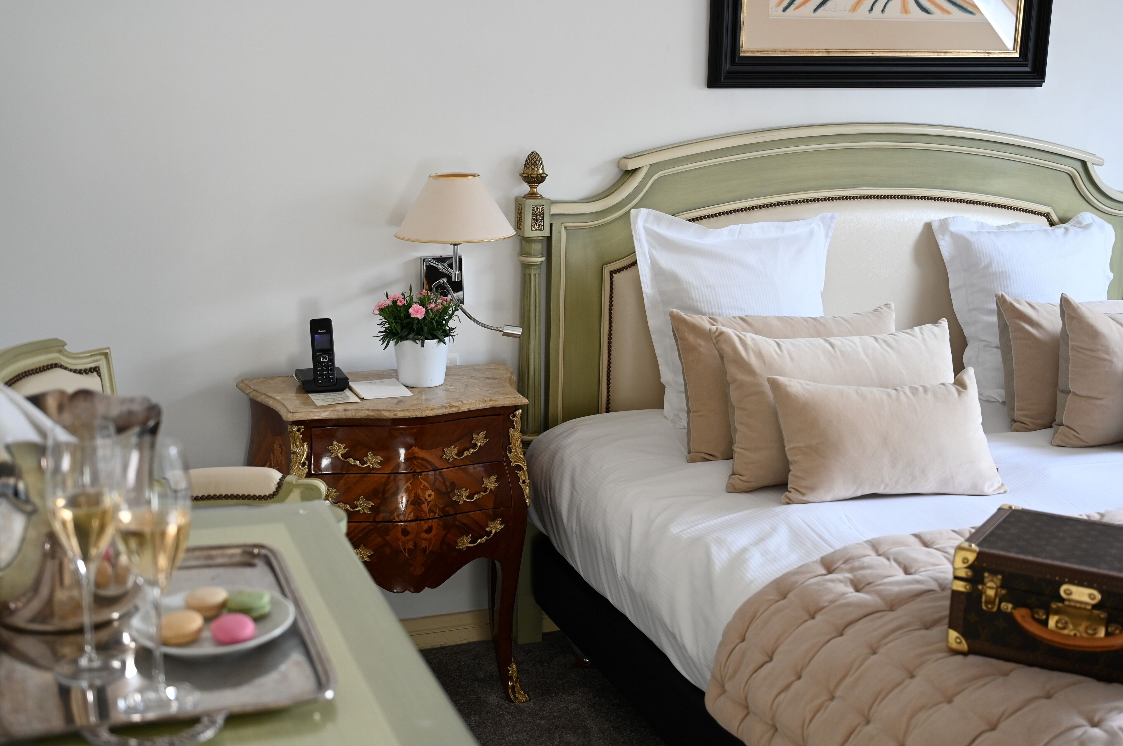 A neatly made bed with white and beige pillows, a bedside table with a lamp and a vase of flowers, and a tray with glasses and macarons.