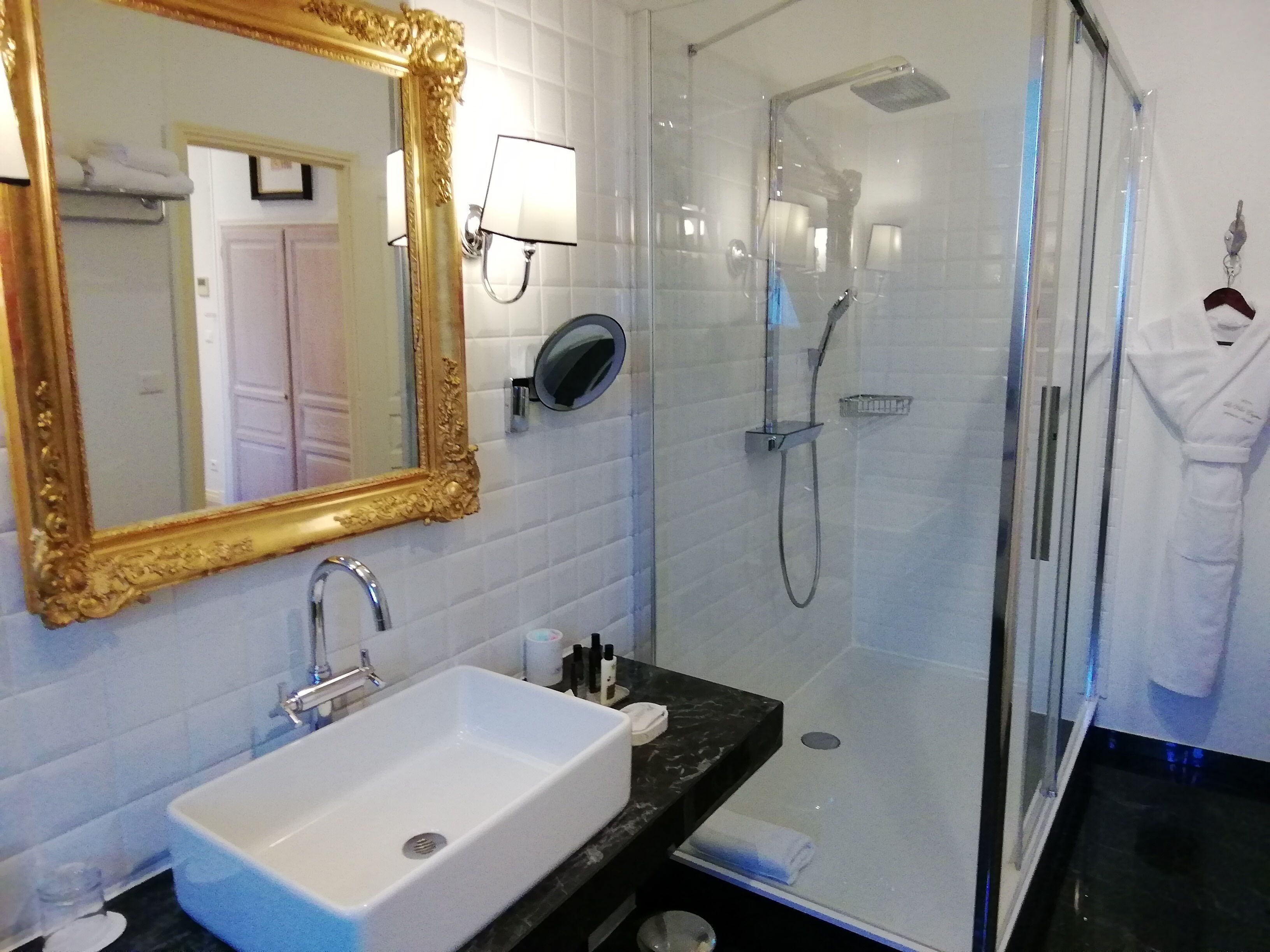 A bathroom with a white sink, a gold-framed mirror, a glass-enclosed shower, and a white robe hanging on the wall.