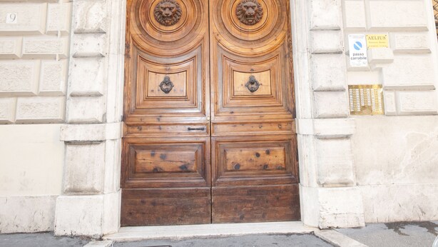 Uma porta dupla de madeira com esculturas elaboradas, em um edifício com fachada de pedra e uma pequena varanda acima.