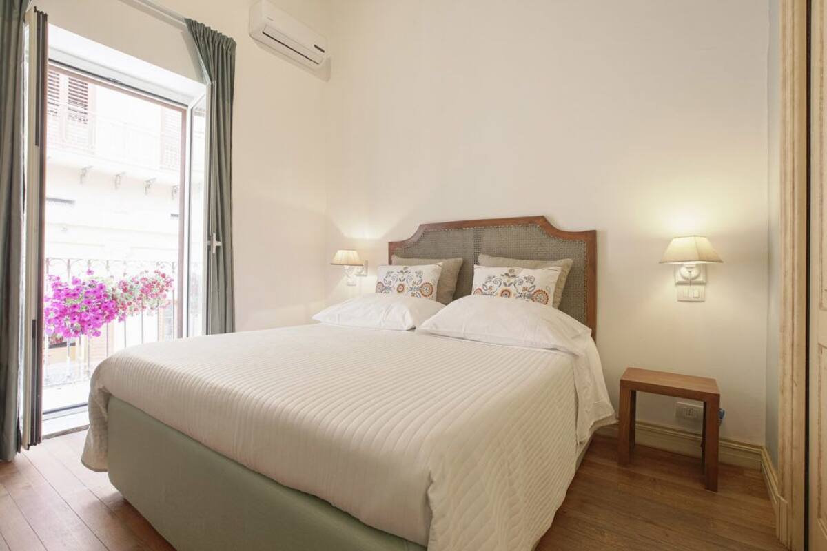 A bedroom with a large bed, two bedside lamps, a small bedside table, and a window with curtains.