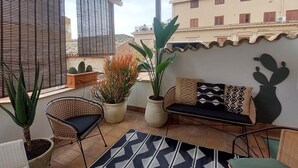 A covered patio area with a seating arrangement, potted plants, and a view of buildings in the background. - Camere a Sud (Agrigento)