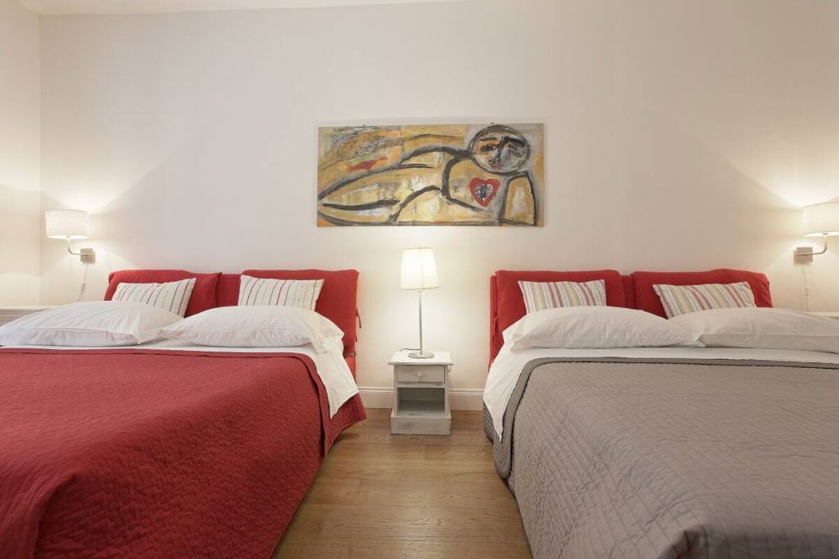 A hotel room with two beds, each with red and white bedding, a small bedside table with a lamp, and a painting on the wall.