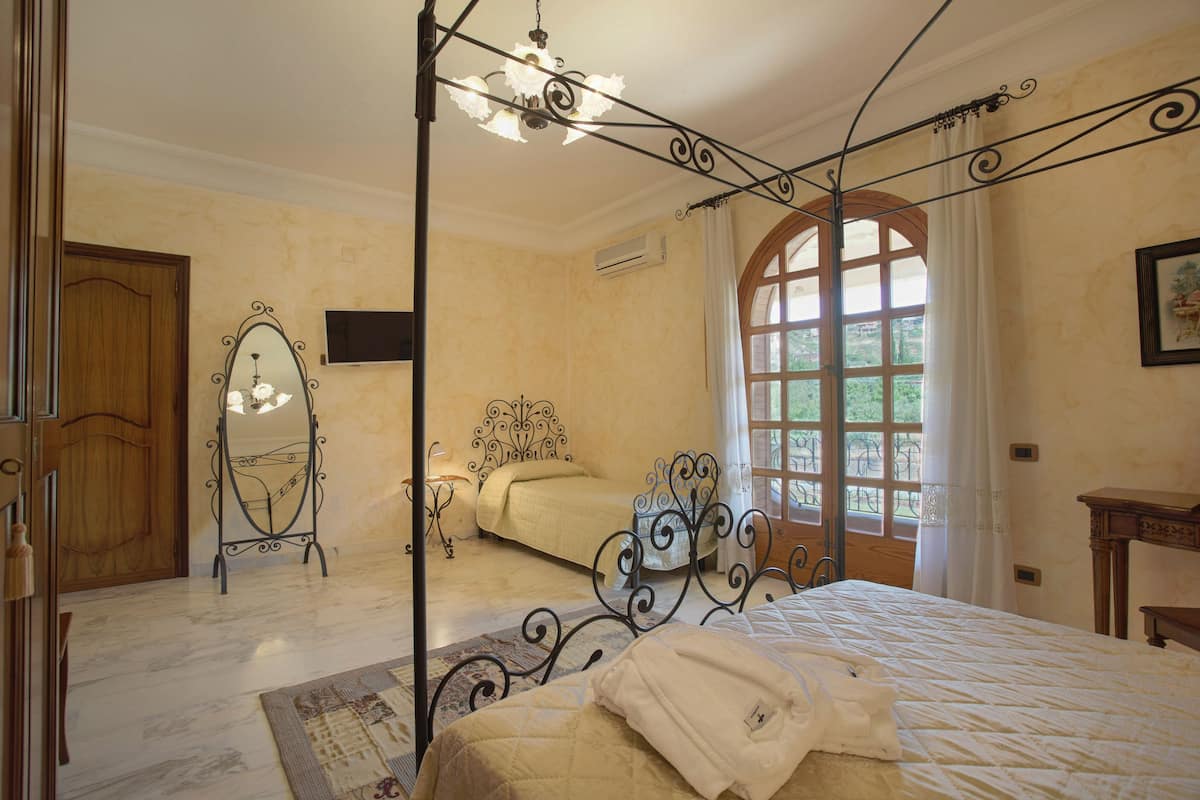 A bedroom with a four-poster bed, a wooden door, a mirror, and a large window with curtains.