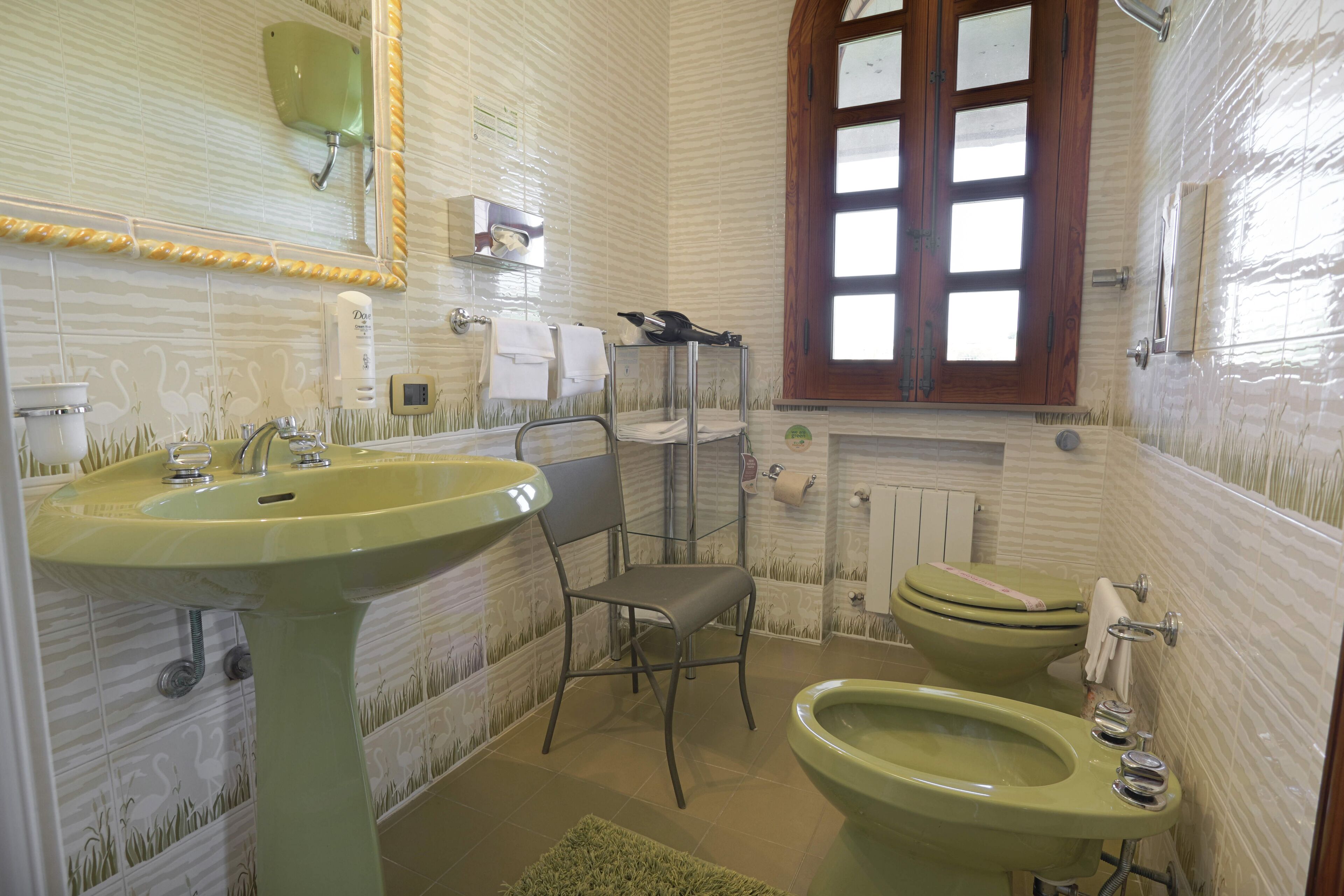 Un bagno con un lavandino verde, un water e una sedia. Si vede una finestra con stipiti in legno e uno specchio sopra il lavandino.