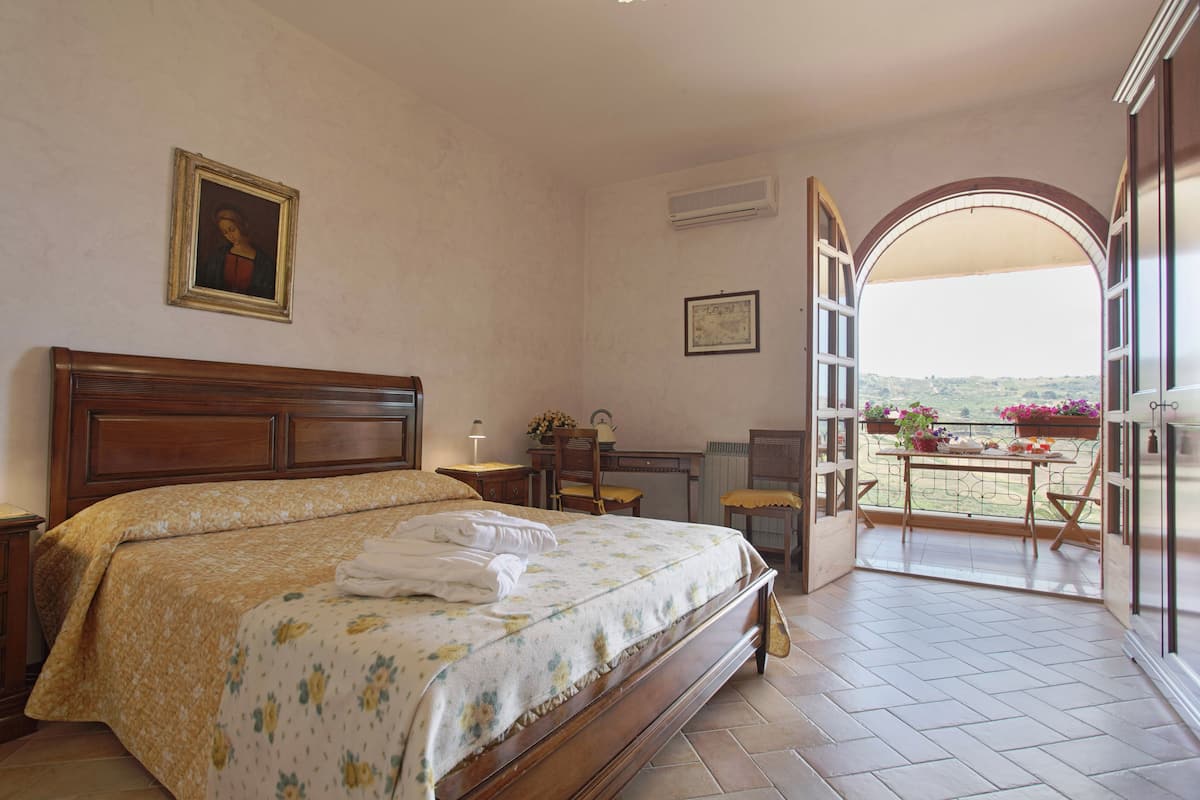 A bedroom with a large bed, a desk, and a view of a balcony with flowers.