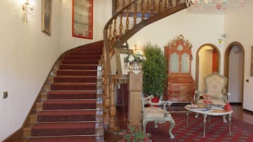 Una imponente escalera con barandilla de madera y peldaños alfombrados de rojo, que conduce a una sala con muebles antiguos, un candelabro y un espejo en la pared.
