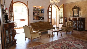 Un salon spacieux avec un canapé marron, deux fauteuils, une table basse et un tapis à motifs. On y trouve une bibliothèque remplie de livres et d’objets décoratifs, un meuble en bois avec des portes en verre et un lustre.
