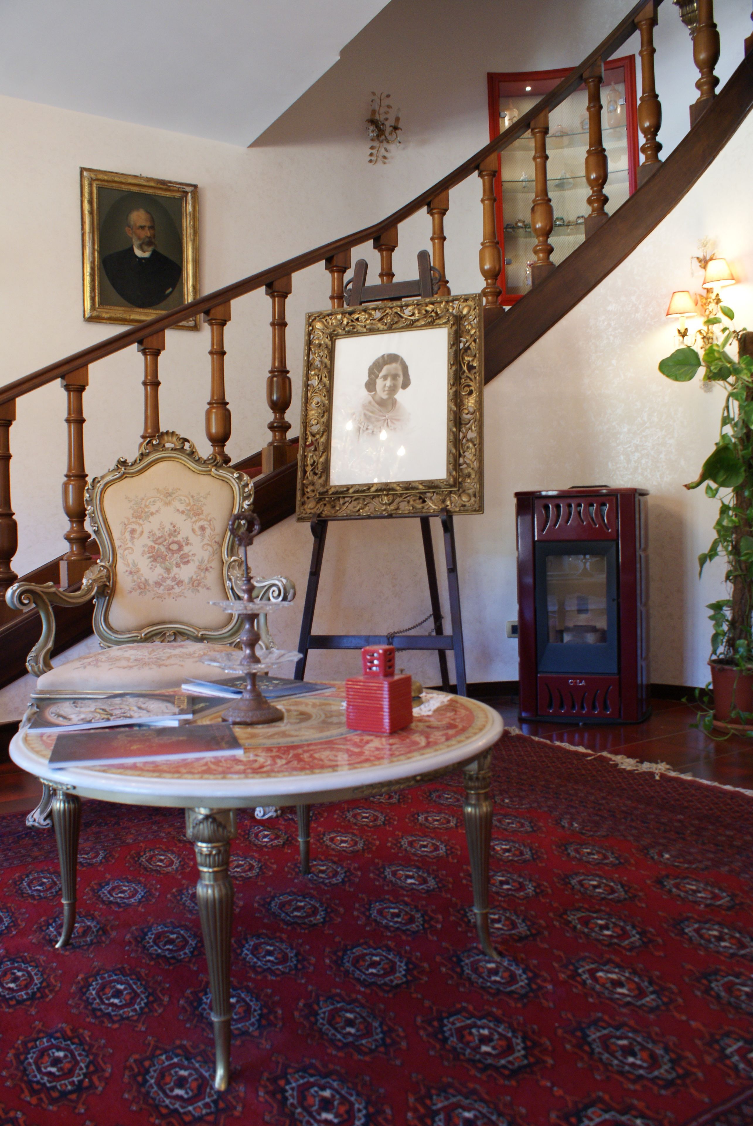 Una escalera con barandas de madera, un retrato enmarcado en la pared, una mesa con mantel rojo y una alfombra estampada.