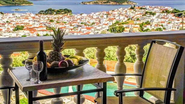 Varanda com mesa posta, garrafa, copos e tigela de frutas, com vista para uma cidade costeira com edifícios de telhado vermelho e vista para o mar.