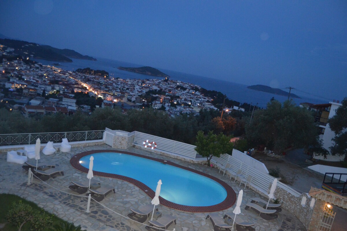 A swimming pool with lounge chairs and umbrellas, surrounded by stone walls and a view of a coastal town at dusk.