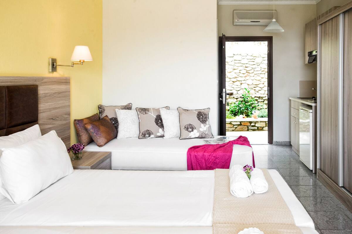 A hotel room with a bed, a sofa, a TV, a lamp, and a view of a stone wall and greenery outside.