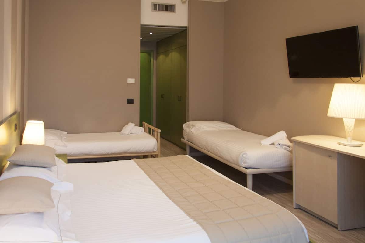 A hotel room with two single beds, a desk with a lamp, a flat-screen TV mounted on the wall, and a green door in the background.