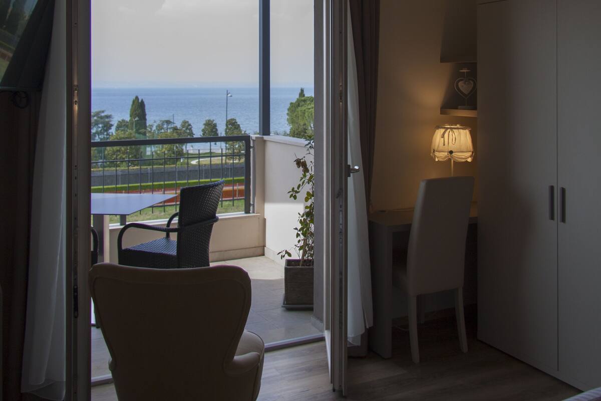 A room with a bed, a desk, and a chair, with a view of the sea and a balcony.