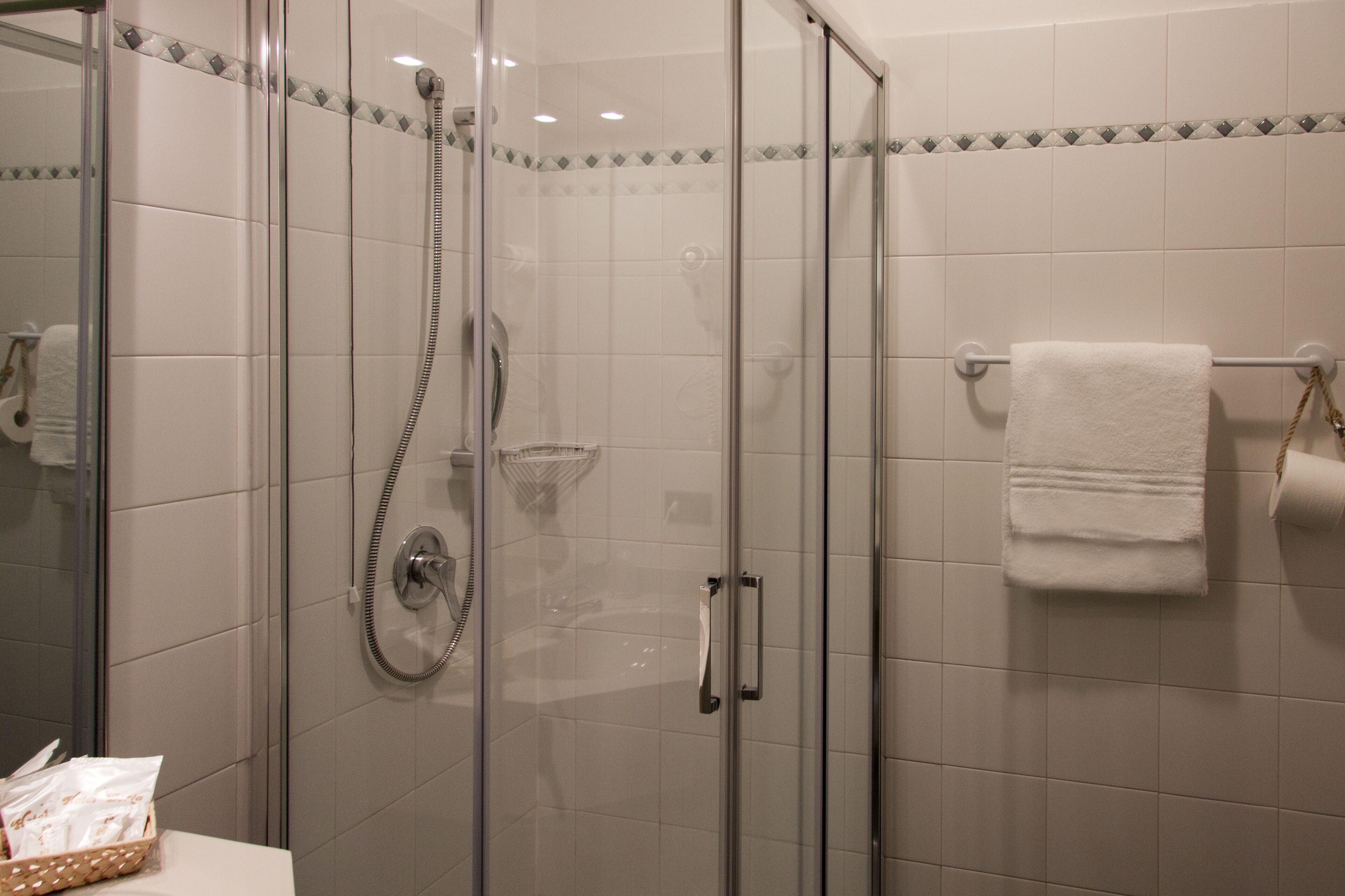 A glass-enclosed shower with a handheld showerhead, a towel rack with a white towel, and a basket of toiletries on the sink.