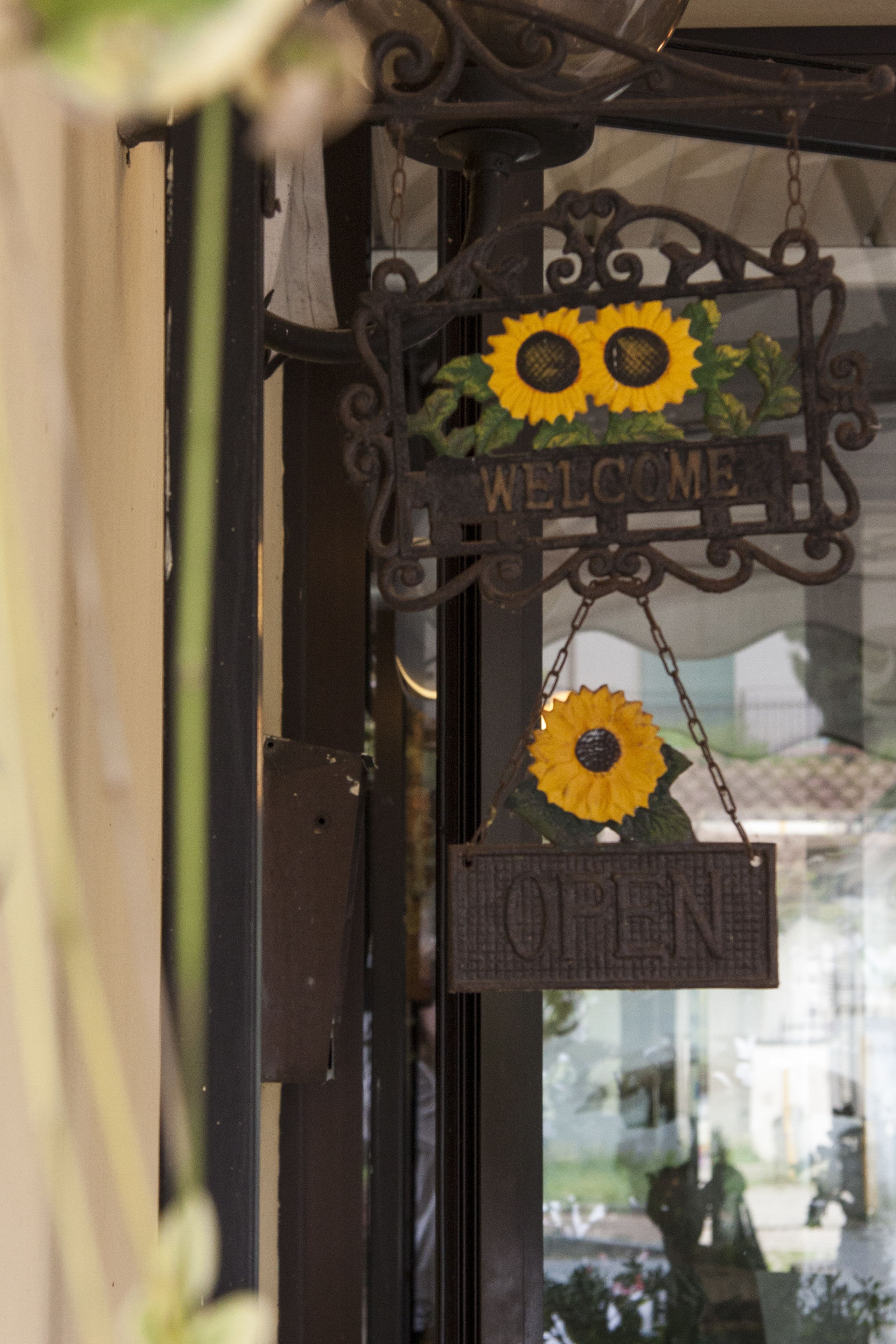 Un letrero de hierro forjado con un girasol y las palabras 'WELCOME' y 'OPEN' colgadas de una puerta.