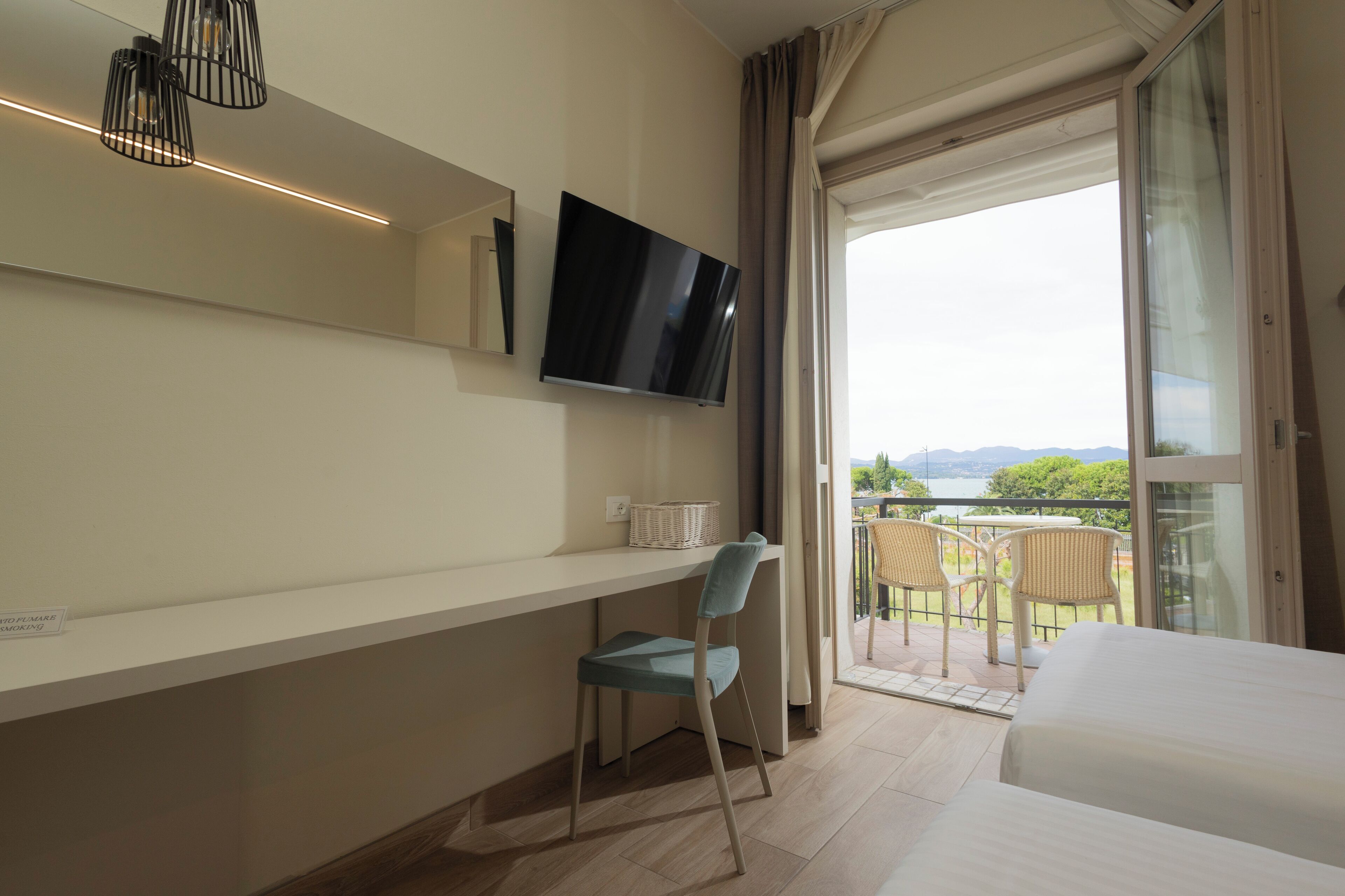A modern hotel room with a large bed, a TV mounted on the wall, a desk with a chair, and a balcony with outdoor seating.