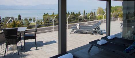 A balcony with sun loungers, a table, and chairs overlooking a body of water and mountains.
