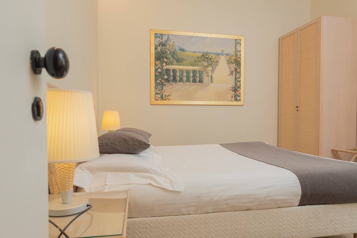 A bedroom with a bed, bedside table, lamp, and a painting on the wall.