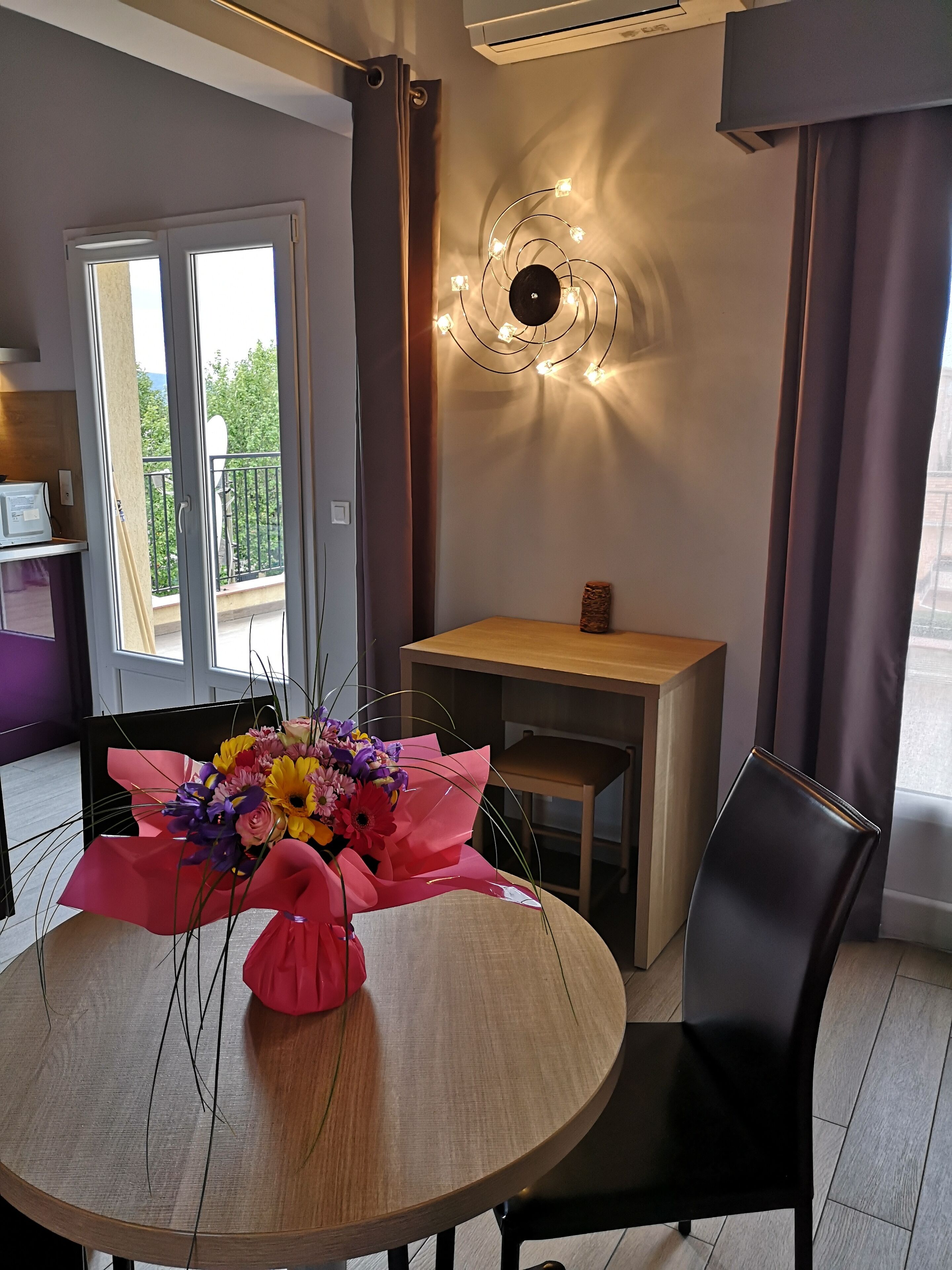 Una habitación moderna con una mesa redonda de madera, un ramo de flores, una pequeña mesita de madera, una silla negra y una lámpara decorativa.