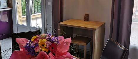 Una habitación moderna con una mesa redonda de madera, un ramo de flores, una pequeña mesita de madera, una silla negra y una lámpara decorativa.