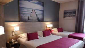 Habitación de hotel con dos camas, cada una con ropa blanca y almohadones rojos, una mesita de noche con lámpara, un escritorio con silla y un florero con flores.
