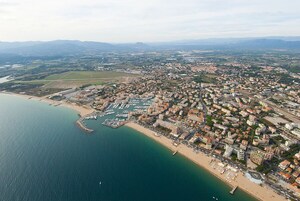 Vista aérea de una ciudad costera con un puerto deportivo, zona urbana densa y campos verdes circundantes.