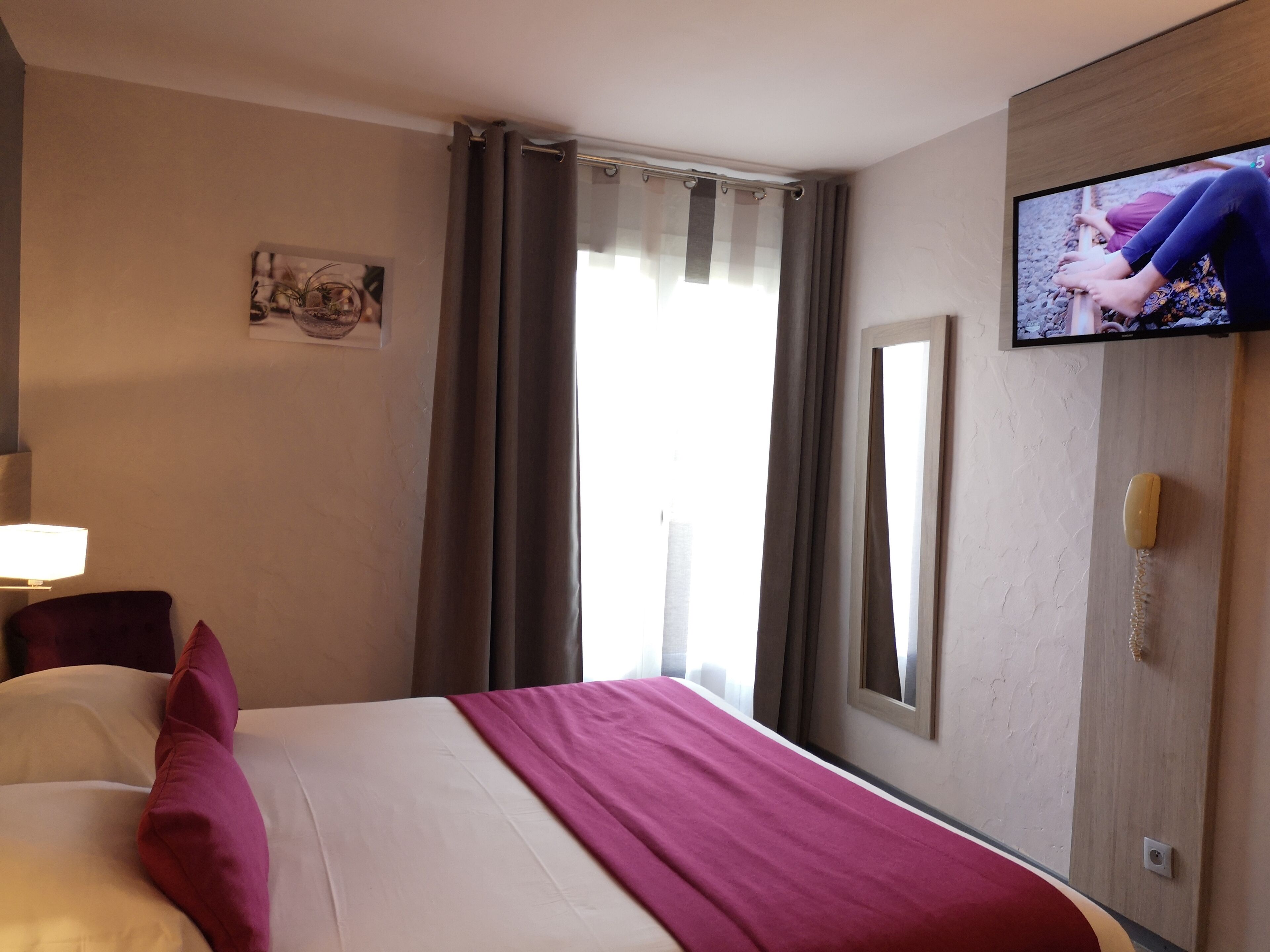A hotel room with a bed, a television, a window with curtains, and a wall-mounted picture.