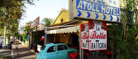 A vintage blue car parked in front of a building with a sign reading "Hotel Atoll Hotel" and another sign for "Be Bella Trattoria".