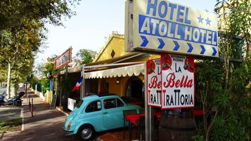 A vintage blue car parked in front of a building with a sign reading "Hotel Atoll Hotel" and another sign for "Be Bella Trattoria".