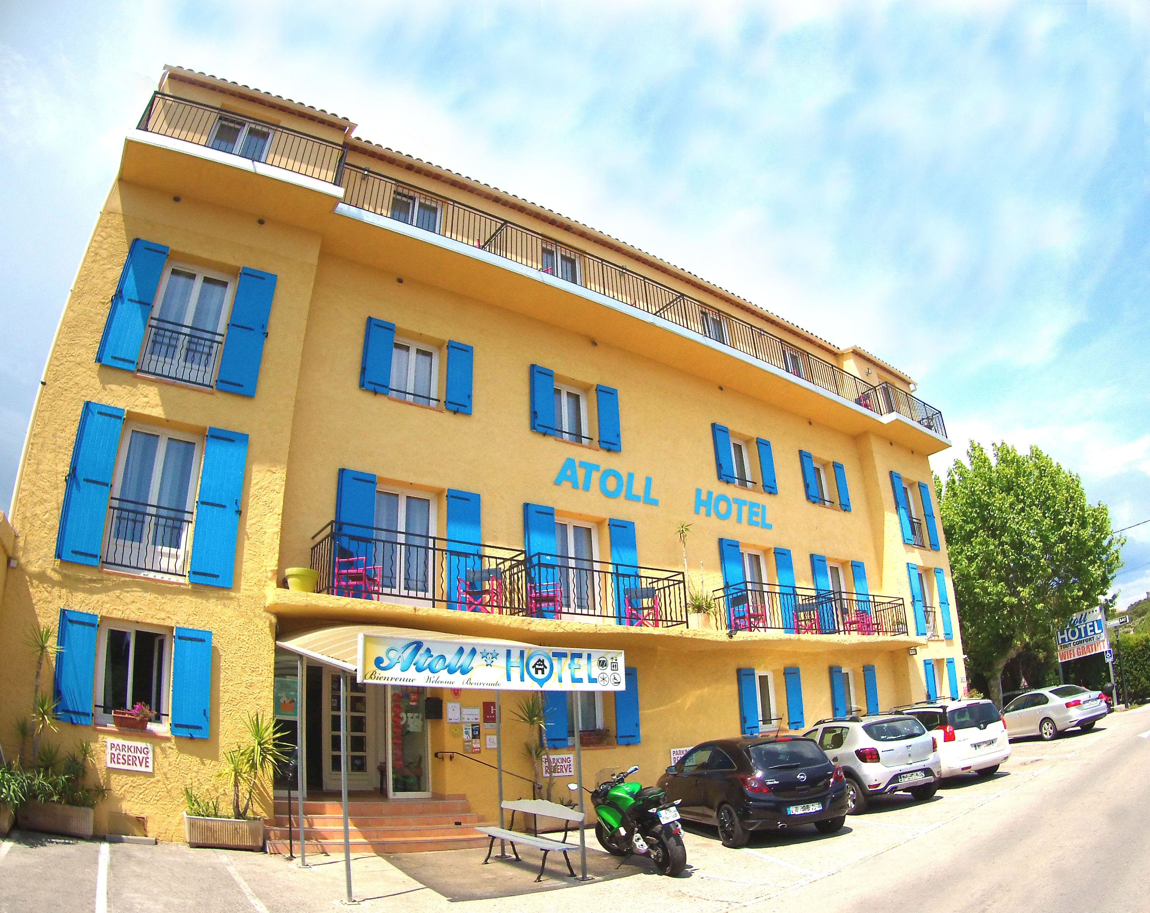 Un edificio giallo con persiane blu e balconi, con la scritta "Atoll Hotel". Sono parcheggiate auto davanti e si vede anche un motociclo.