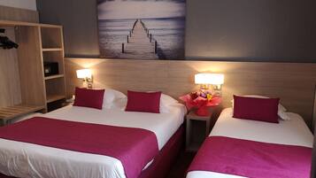 Ein Hotelzimmer mit zwei Betten, jeweils mit weißer Bettwäsche und roten Kissen. Ein Holz-Kopfteil, ein Nachttisch mit Lampe und ein Holzregal sind vorhanden. An der Wand hängt ein Gemälde, das einen Pier zeigt, der ins Meer führt.