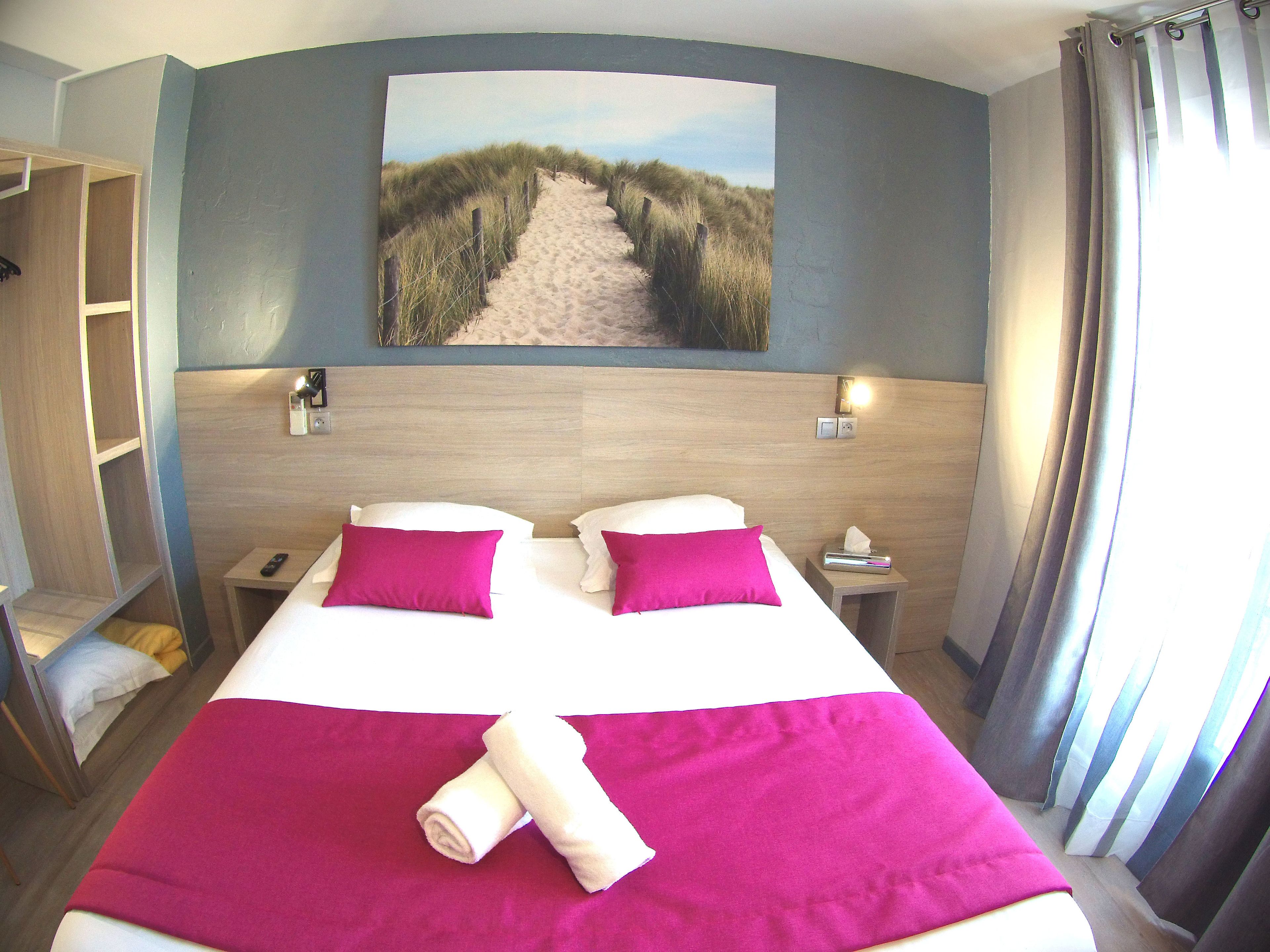 Une chambre d’hôtel avec un lit, deux oreillers, une table de chevet, une télécommande, un tableau représentant un sentier de plage et une fenêtre avec des rideaux.