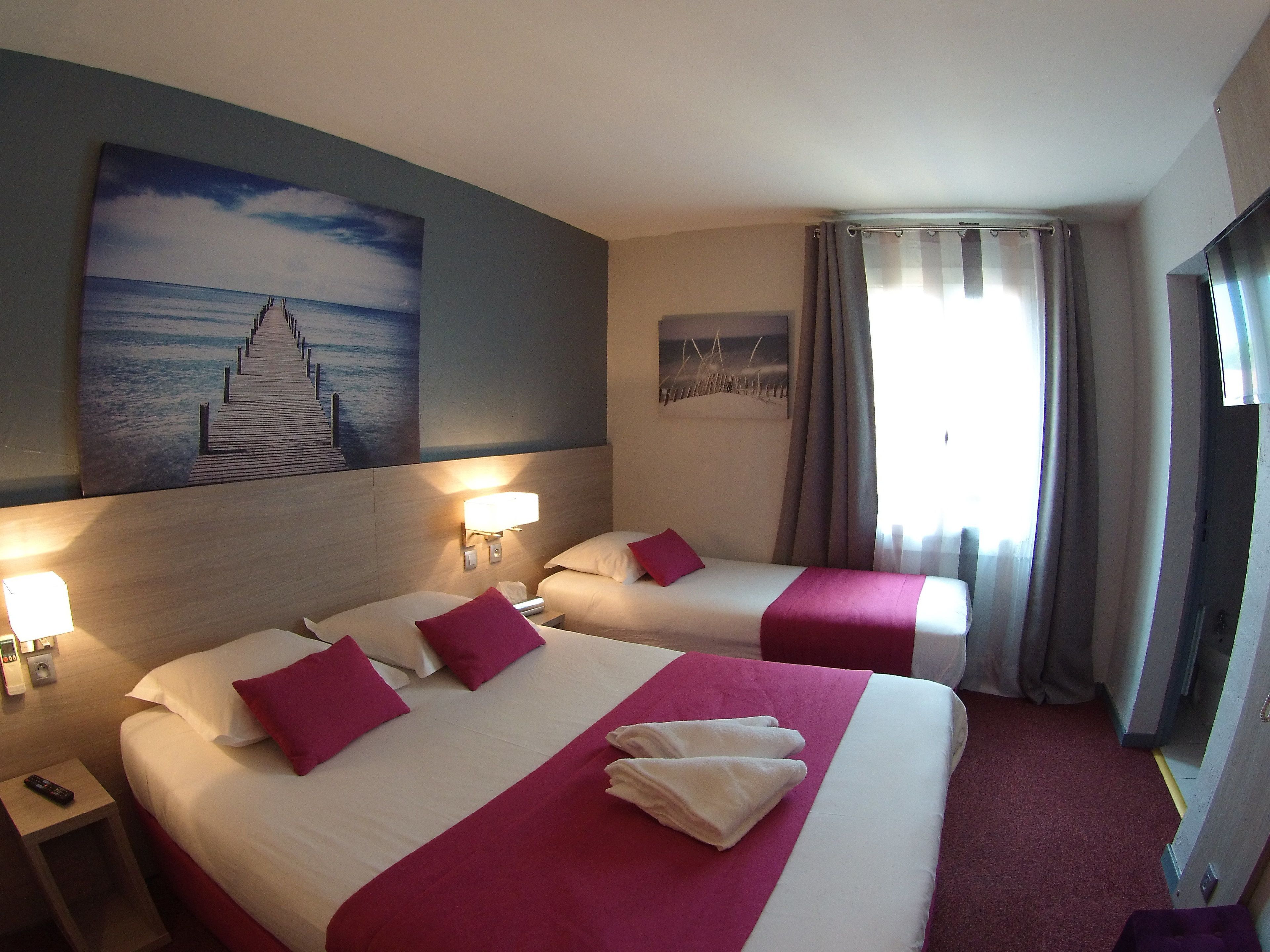 A hotel room with two beds, each with white linens and red accents, a painting of a pier on the wall, a nightstand with a lamp, and a TV on the right.