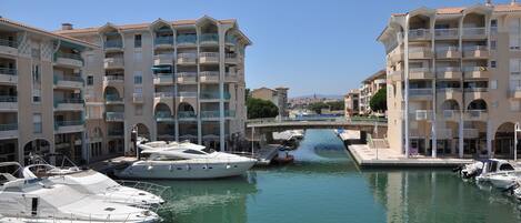 A marina with several boats docked, surrounded by multi-story buildings with balconies and a bridge in the background.