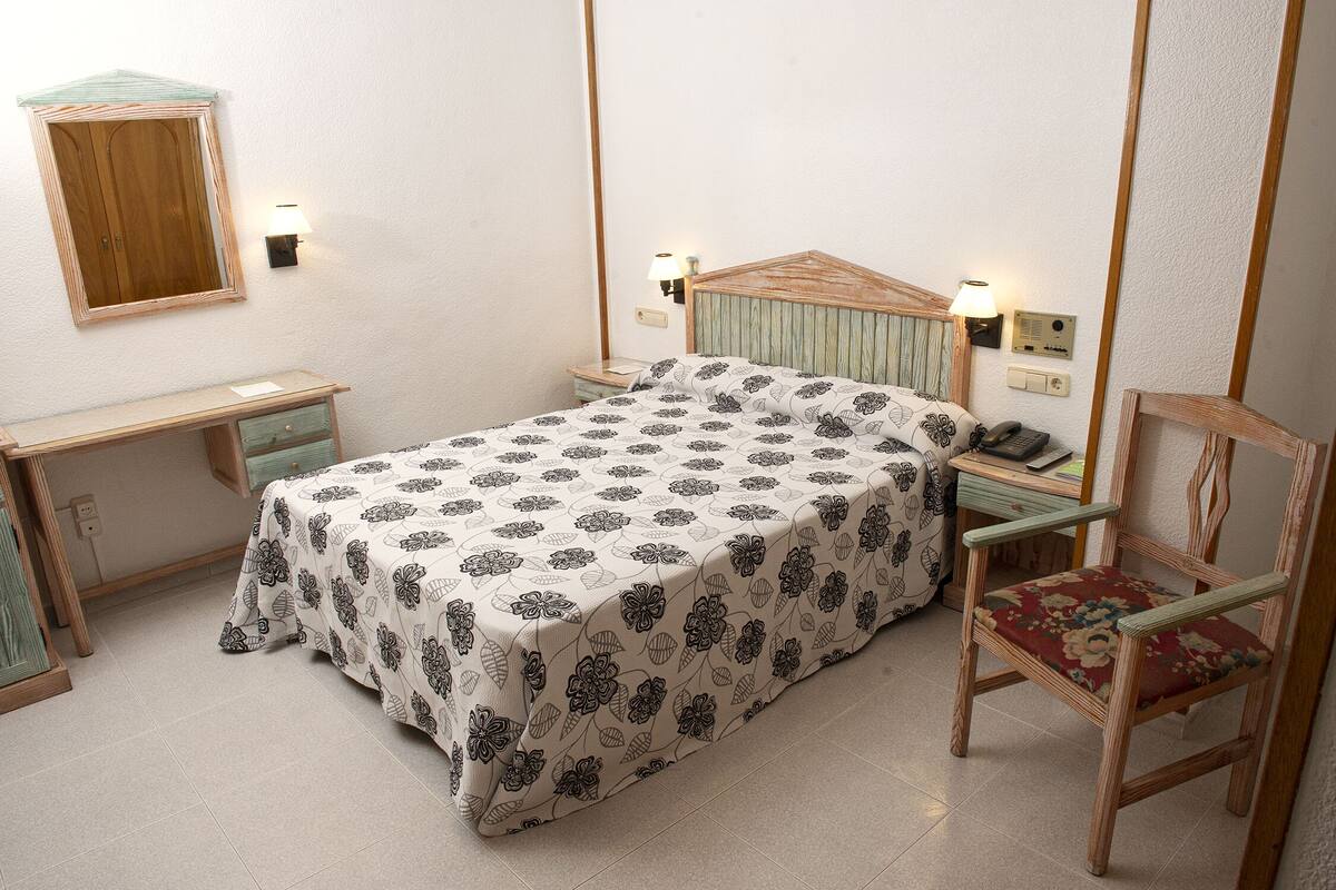Un dormitorio con cama, mesita de noche, silla y espejo en la pared.