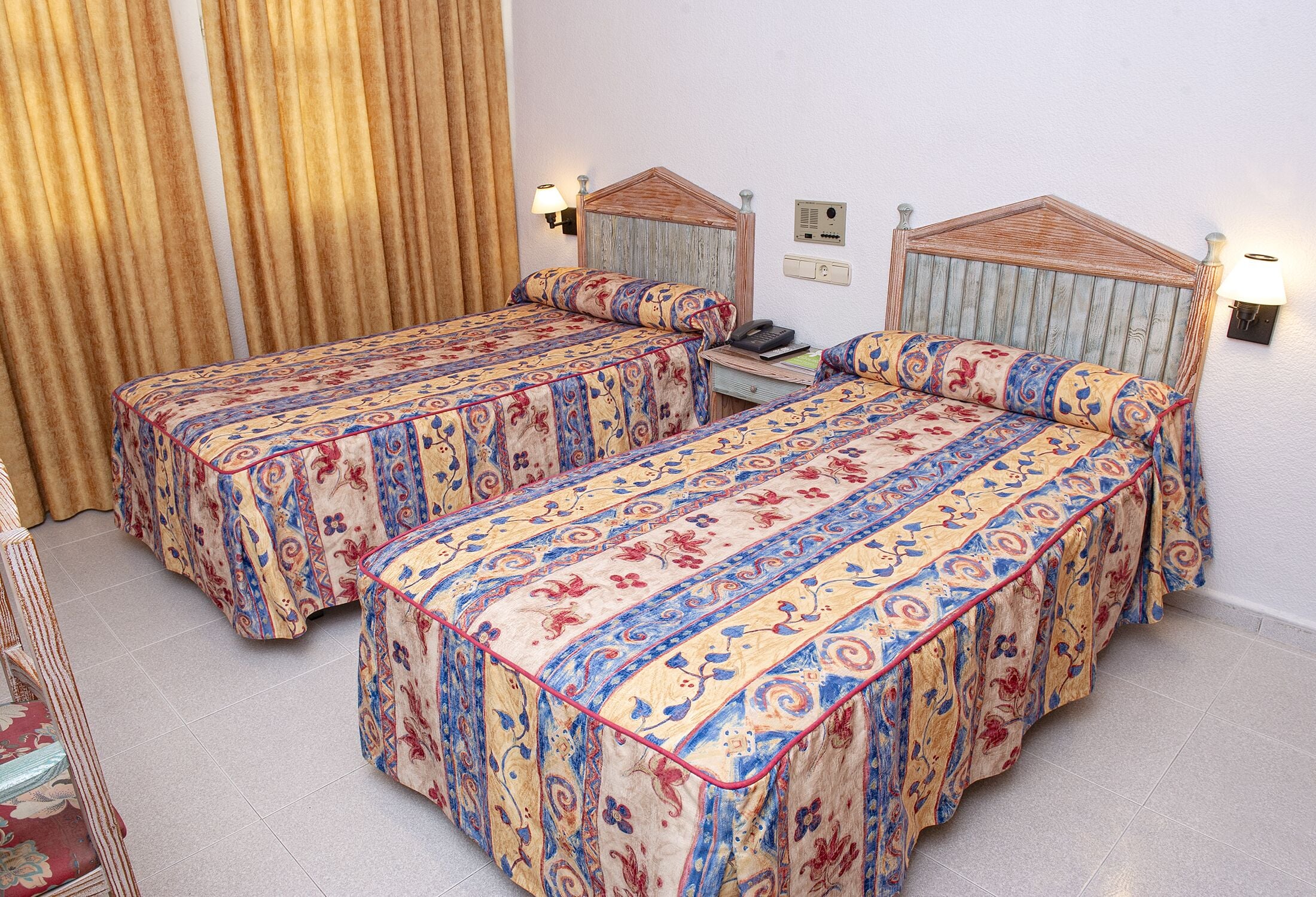A hotel room with two beds, each with a patterned bedspread and headboard, a bedside table with a lamp, and a wall-mounted alarm clock.