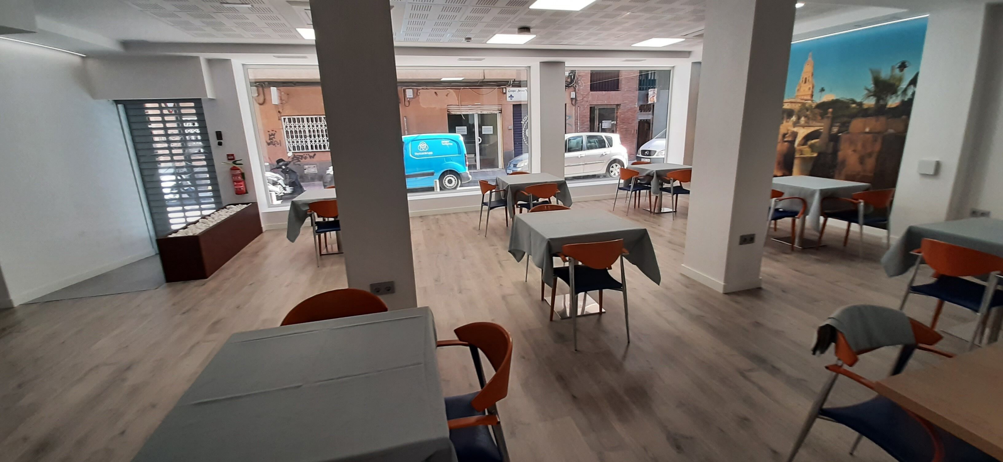 Un espacio interior amplio con varias mesas y sillas, ventanas grandes y vista a la calle.