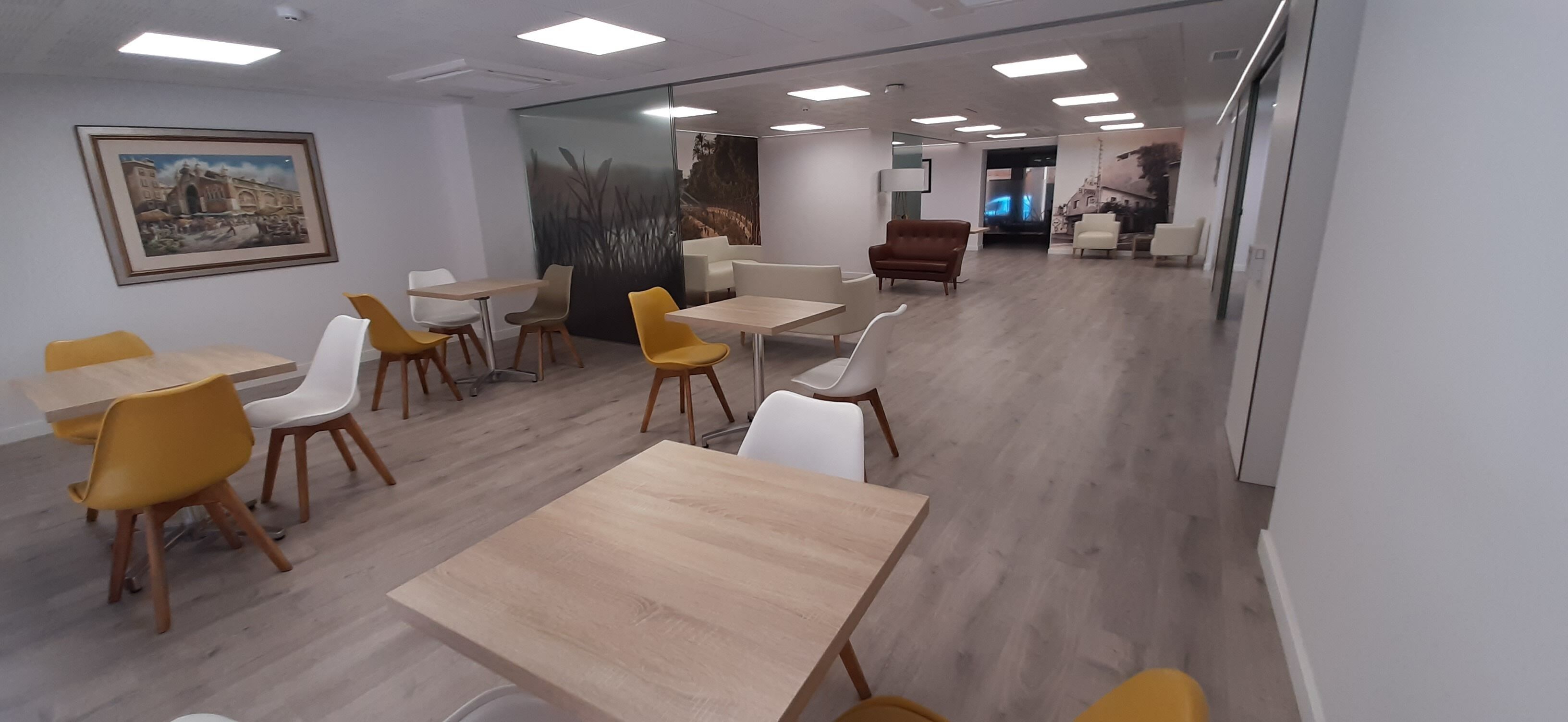 Una sala de reuniones moderna con mesas y sillas de madera, un separador de vidrio y un mural en la pared.