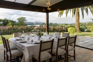 Un espacio exterior para comer con una mesa dispuesta para seis personas, rodeada de sillas, con vistas a un paisaje pintoresco con árboles y una masa de agua.