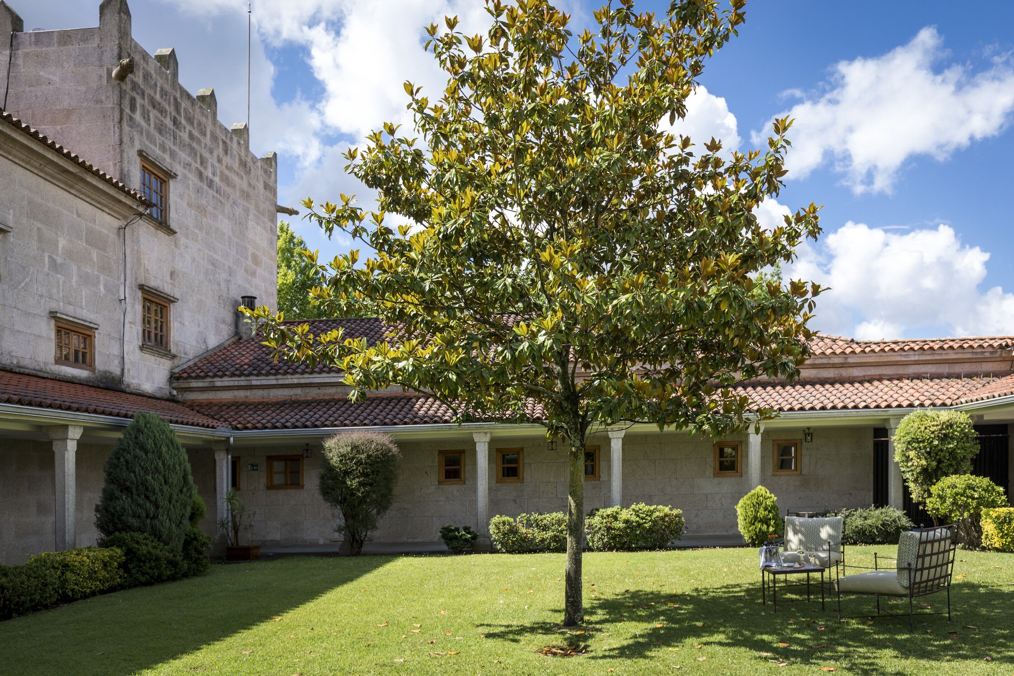 A courtyard with a tree, a covered seating area, and a stone building with a tiled roof.